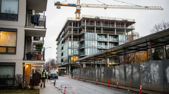 A residential building under construction with a tower crane stands across the street from occupied apartments in a Canadian city neighborhood.