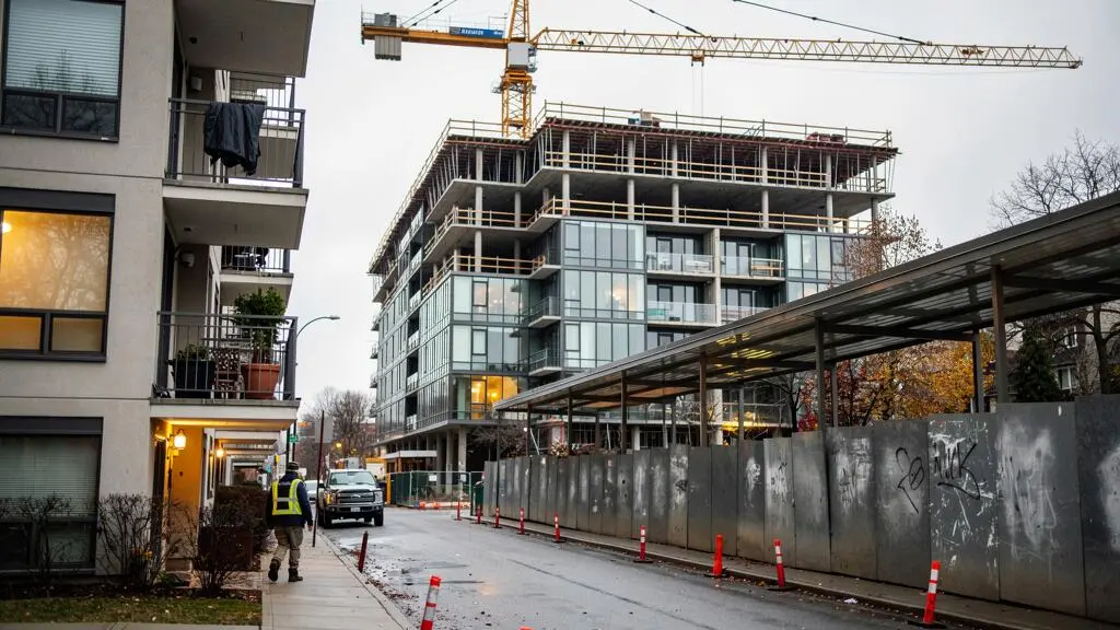 A residential building under construction with a tower crane stands across the street from occupied apartments in a Canadian city neighborhood.